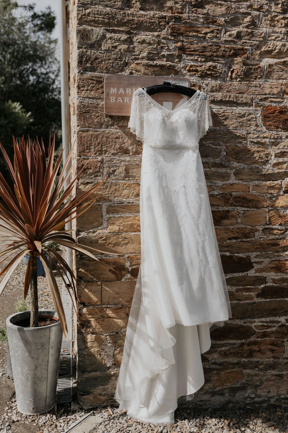 Elegant beaded white wedding dress hanging on stone wall beside tropical plant.