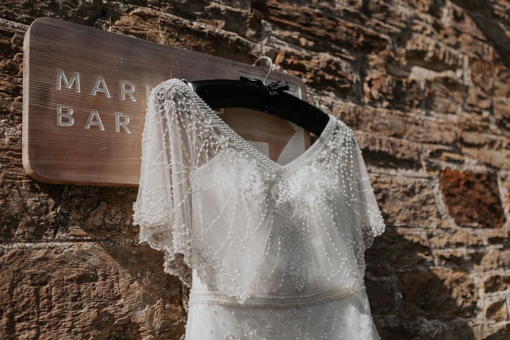 Close-up of beaded white wedding dress with flutter sleeves hanging on stone wall.