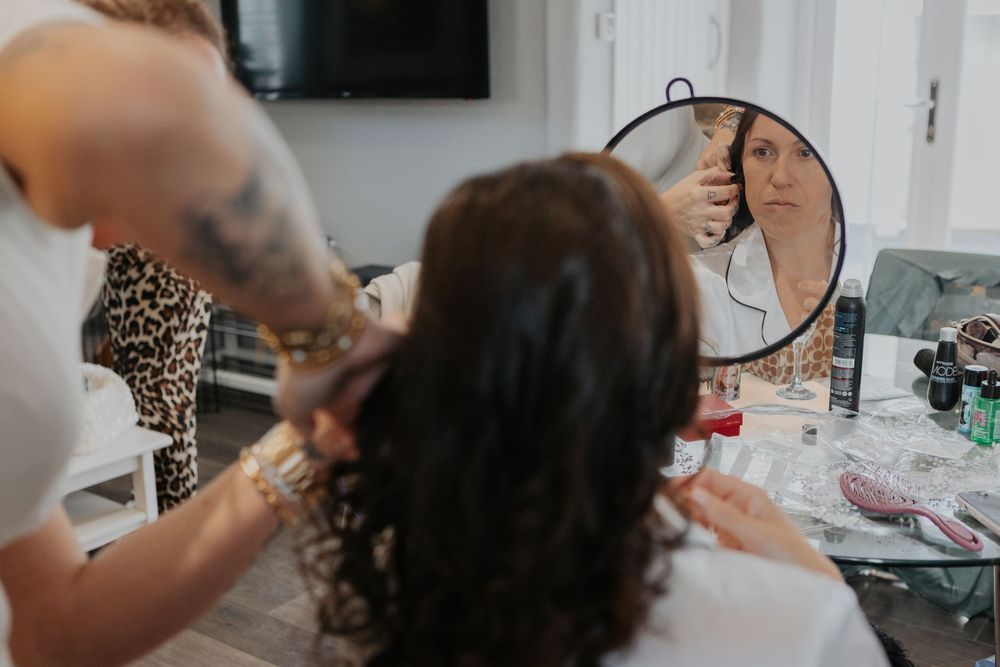 Hairstylist with tattoos styling curly brunette hair while holding a round mirror.