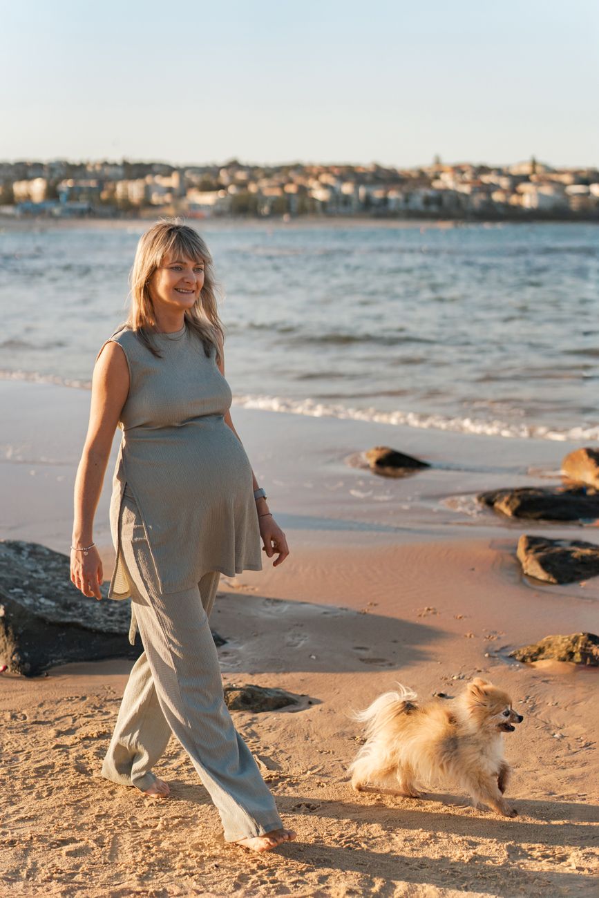 A mom to be in a flowing gray outfit walks along a beach with a golden small dog at sunset during her maternity photoshoot in Sydney