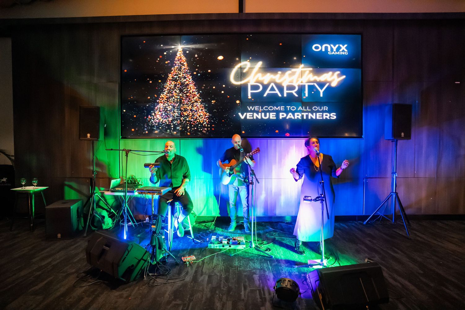 A band performs on stage with colorful lighting at a Christmas party under a digital tree display.