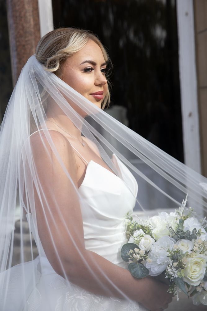 Bride in white wedding dress holds elegant white bouquet while posing with flowing veil in natural light.