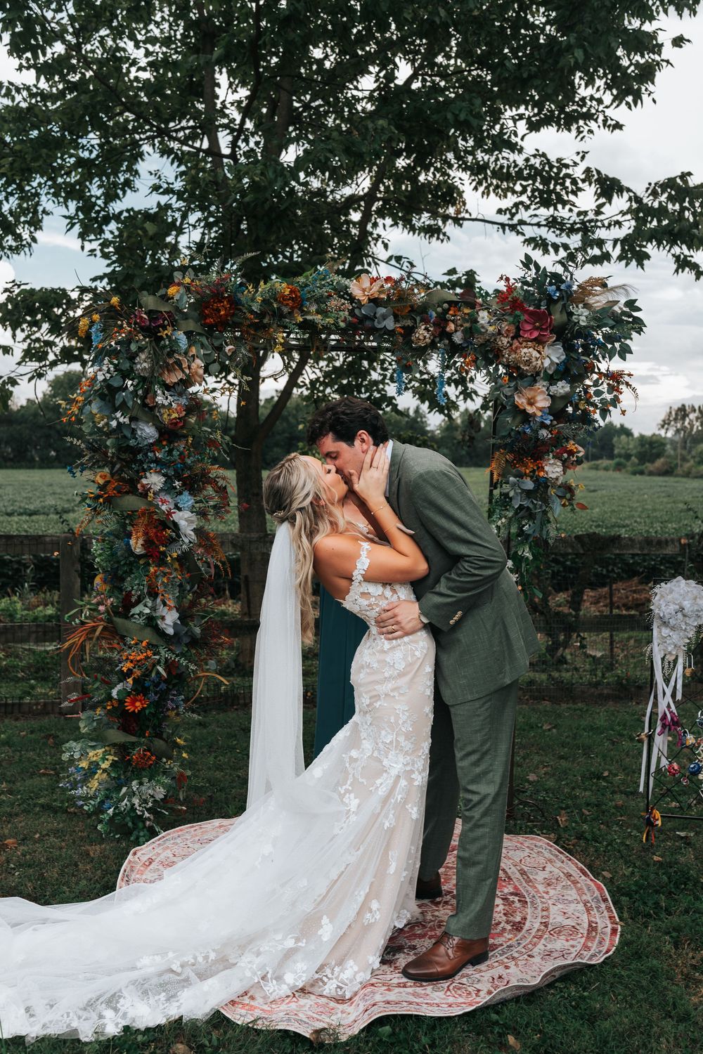 A romantic wedding couple poses under a floral arch with red flowers in an outdoor garden setting.