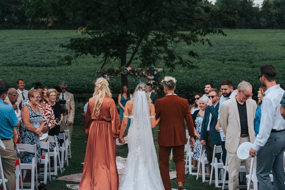 Outdoor garden wedding ceremony setup with white chairs and floral arrangements under tall trees.
