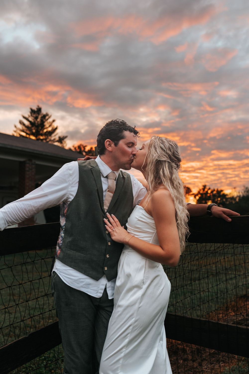 A couple shares romantic kisses at sunset against a dramatic orange and purple sky with a rustic fence in the background.