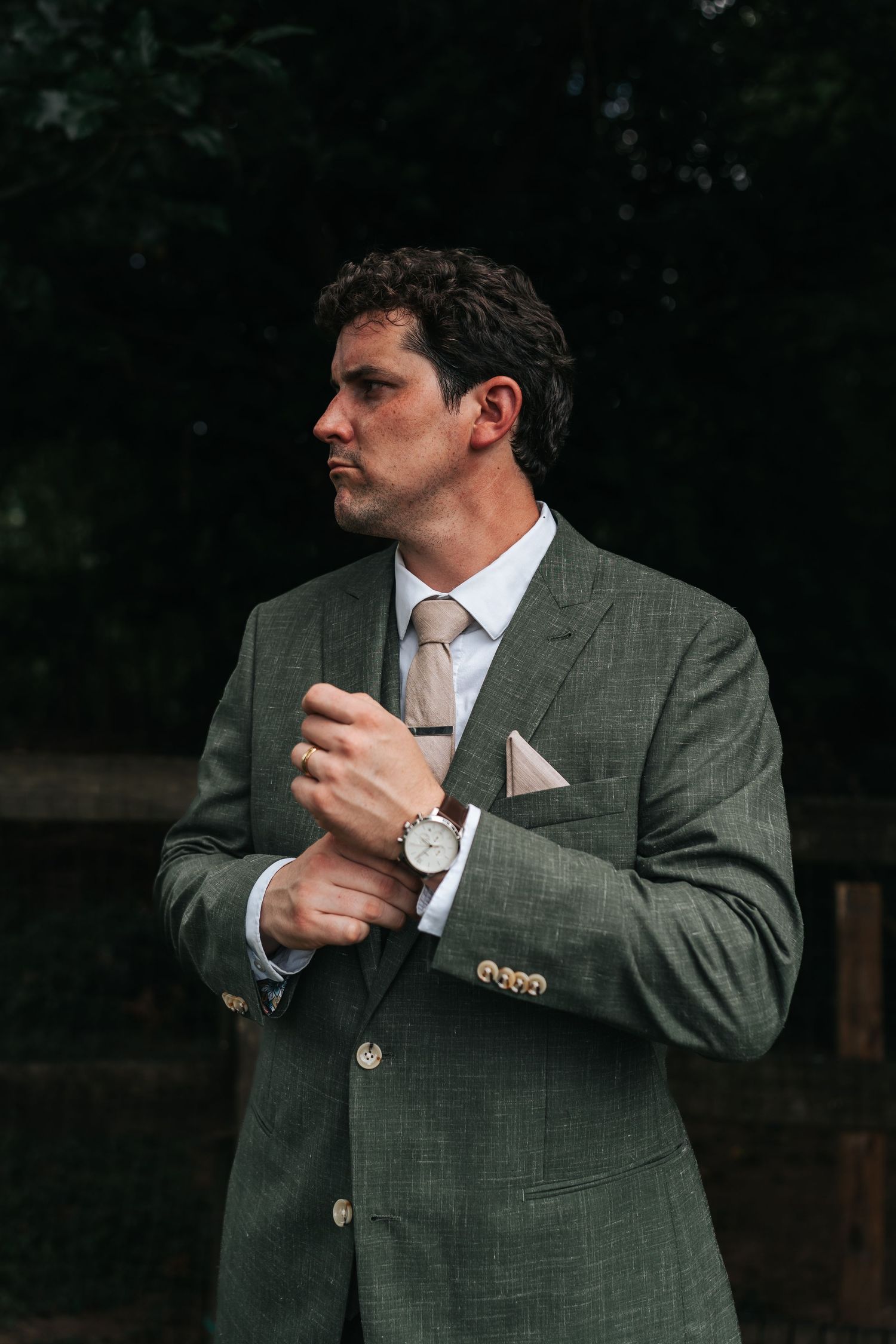 A person wearing a gray suit jacket and tie adjusts their watch while looking to the side in an outdoor setting.