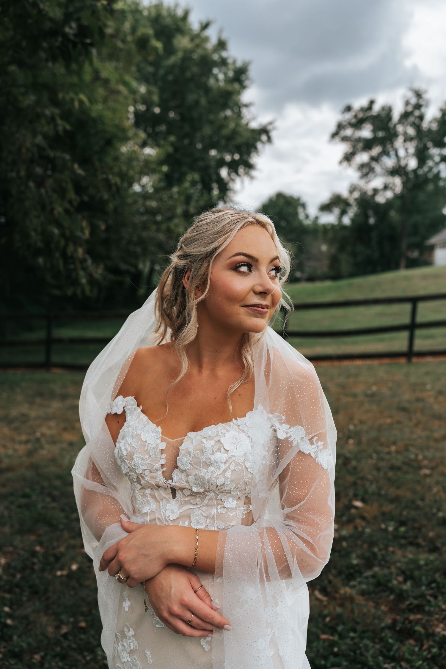 A bride in a white lace wedding dress with sheer sleeves poses outdoors on an overcast day.