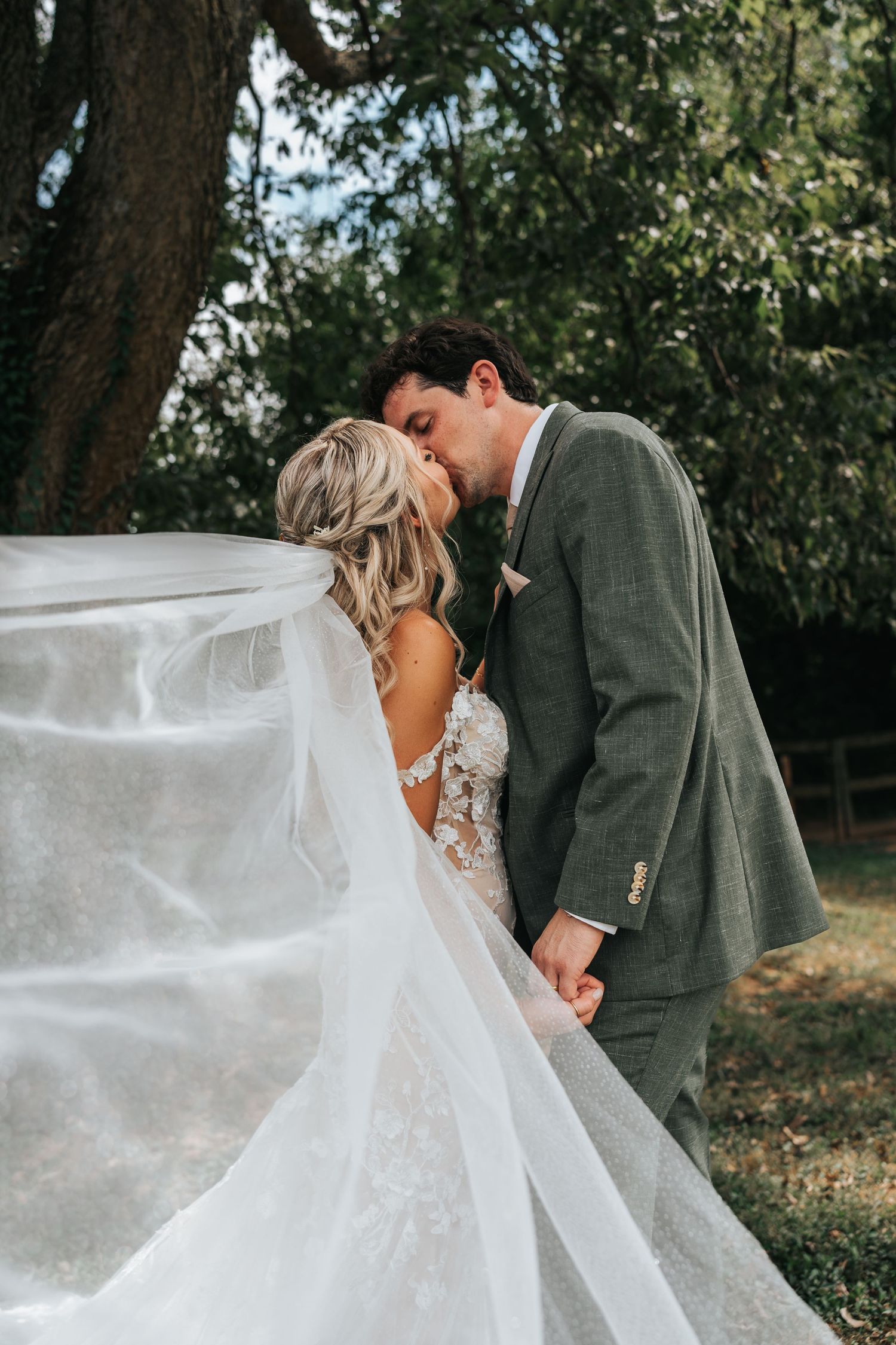A romantic wedding couple embraces outdoors with the bride's long white veil flowing in the breeze.