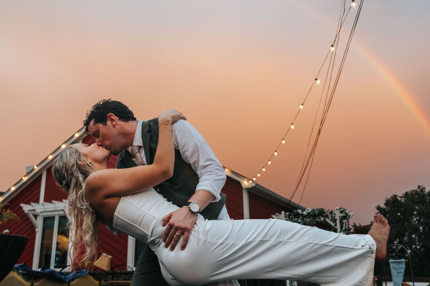 A couple dances and dips during a romantic sunset wedding reception under twinkling string lights.