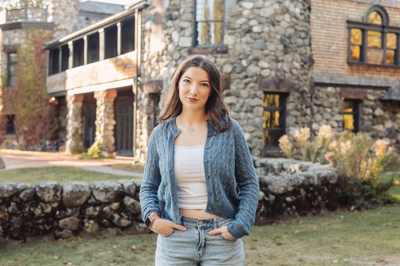 A person in a blue cardigan and jeans poses casually against a rustic stone wall backdrop in autumn.