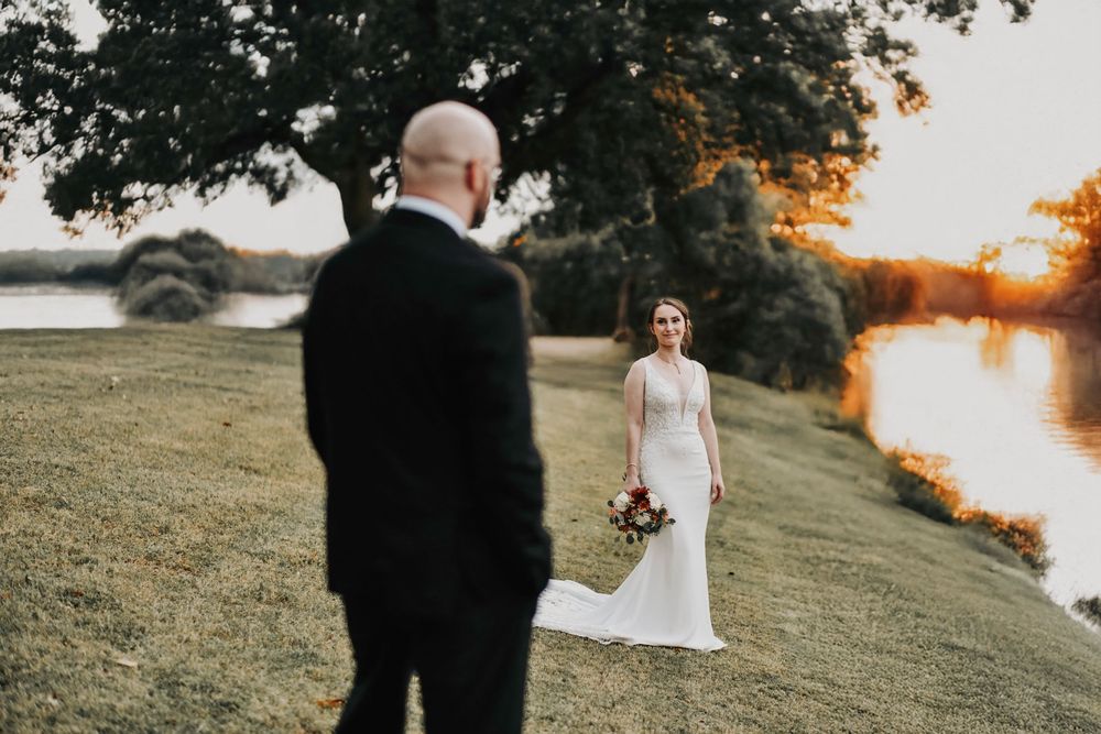 a bride stands a the river's edge and smiles at her husband who stares at her in the foreground.