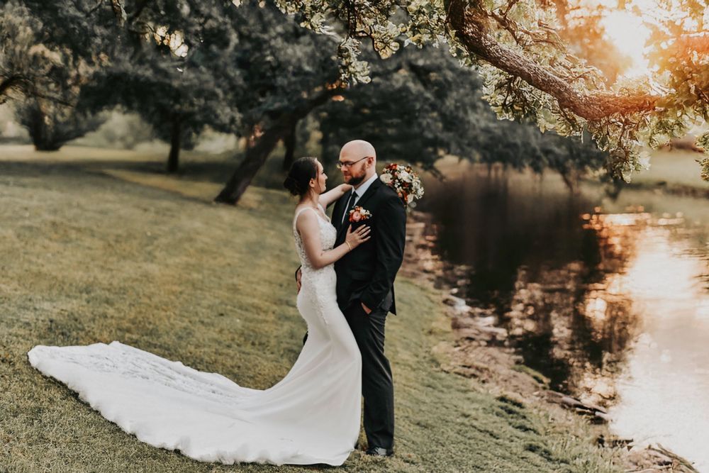 a bride in a modern lace wedding gown embraces her husband as they stand in the sunset on the river bank of her wedding venue in weatherford