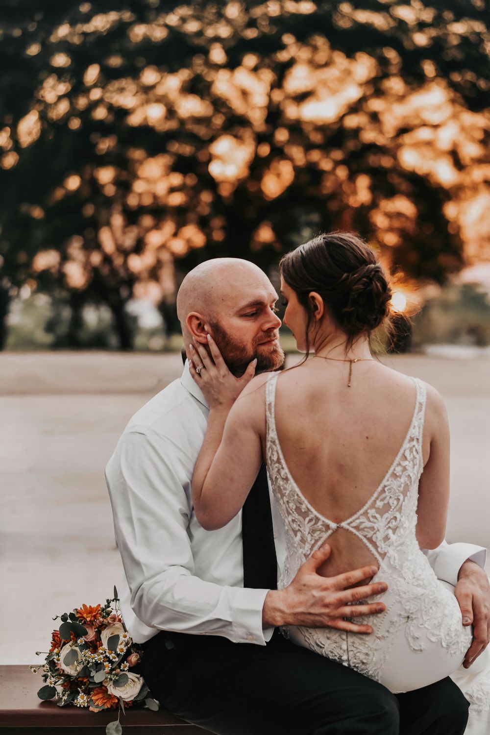 an intimate moment of a bride sitting on her husbands lap as they kiss in the sunlight