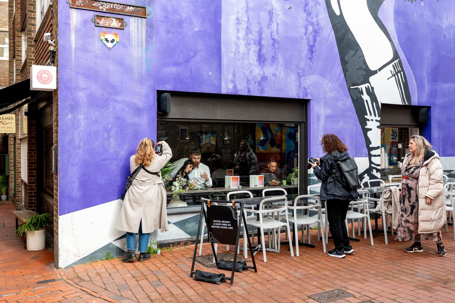 People gather outside a cafe with outdoor seating against a vibrant purple wall in an urban setting.