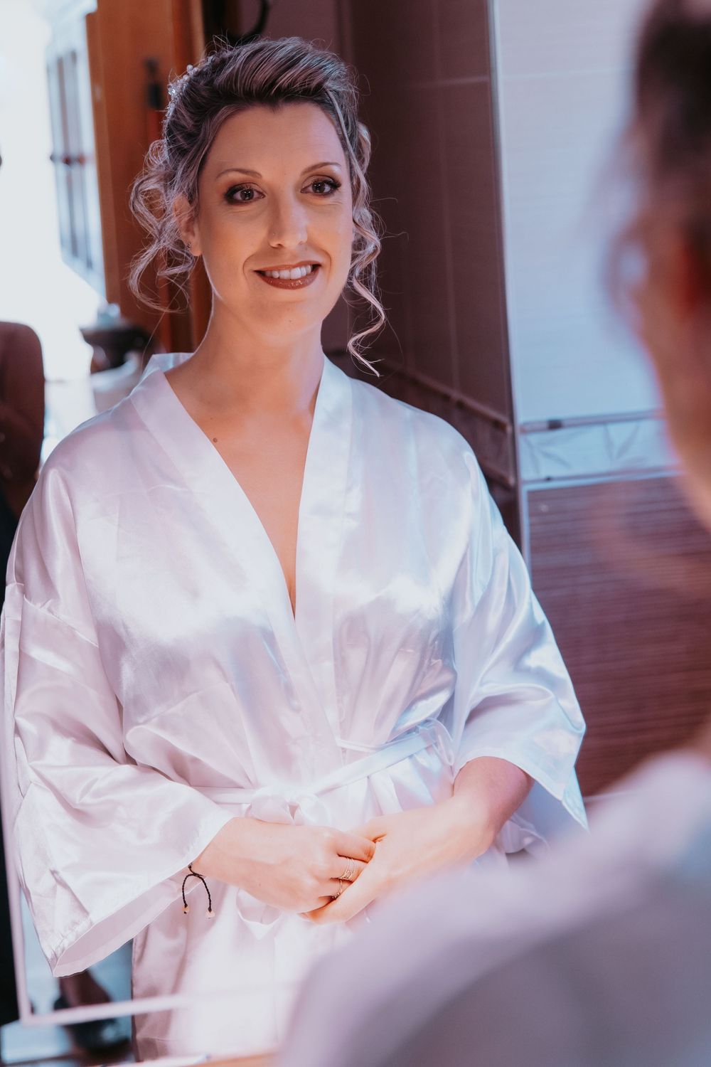 A bride in a white silk robe smiles while getting ready on her wedding day.