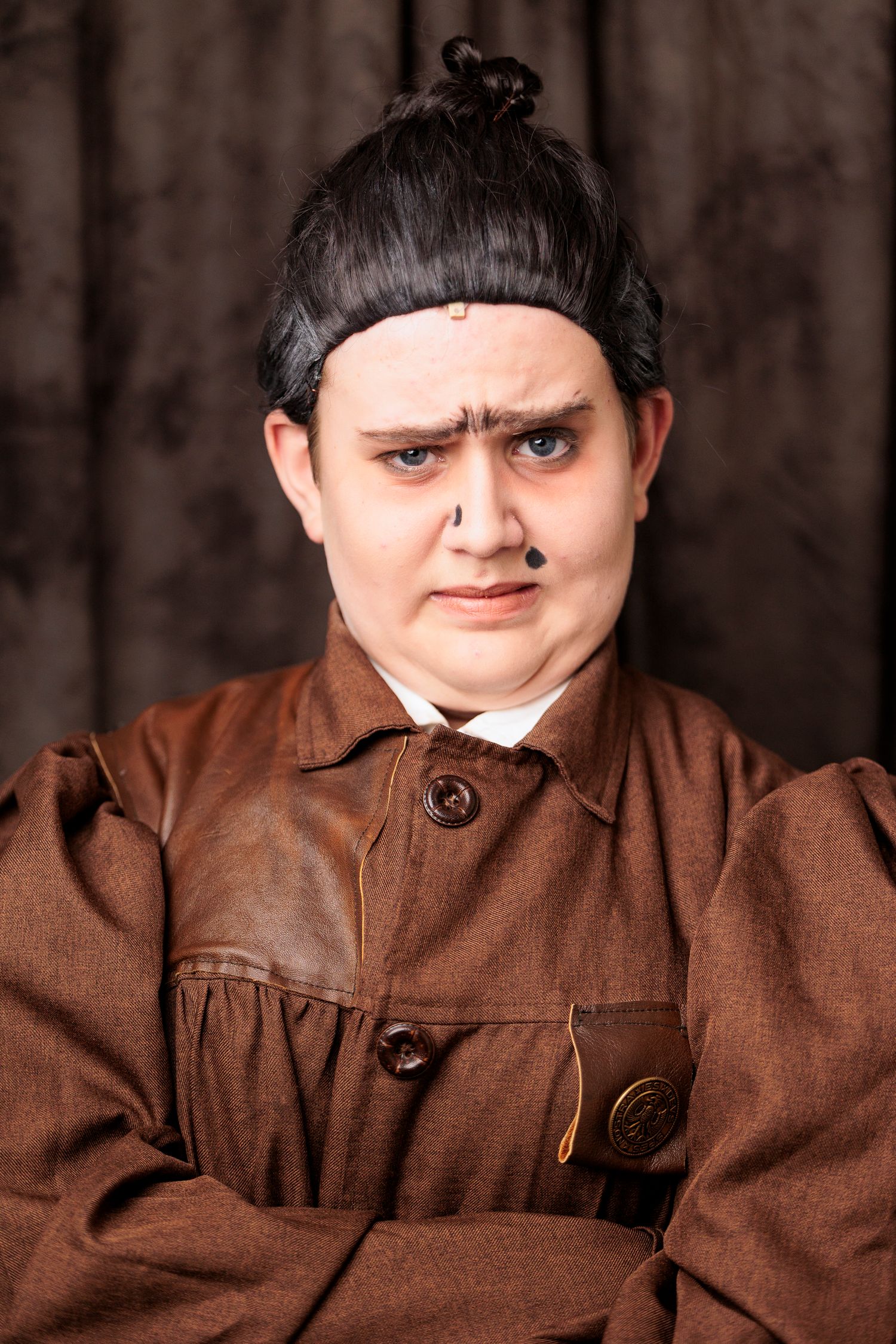 A young performer in a brown costume and black wig portrays a stern character in a stage production.