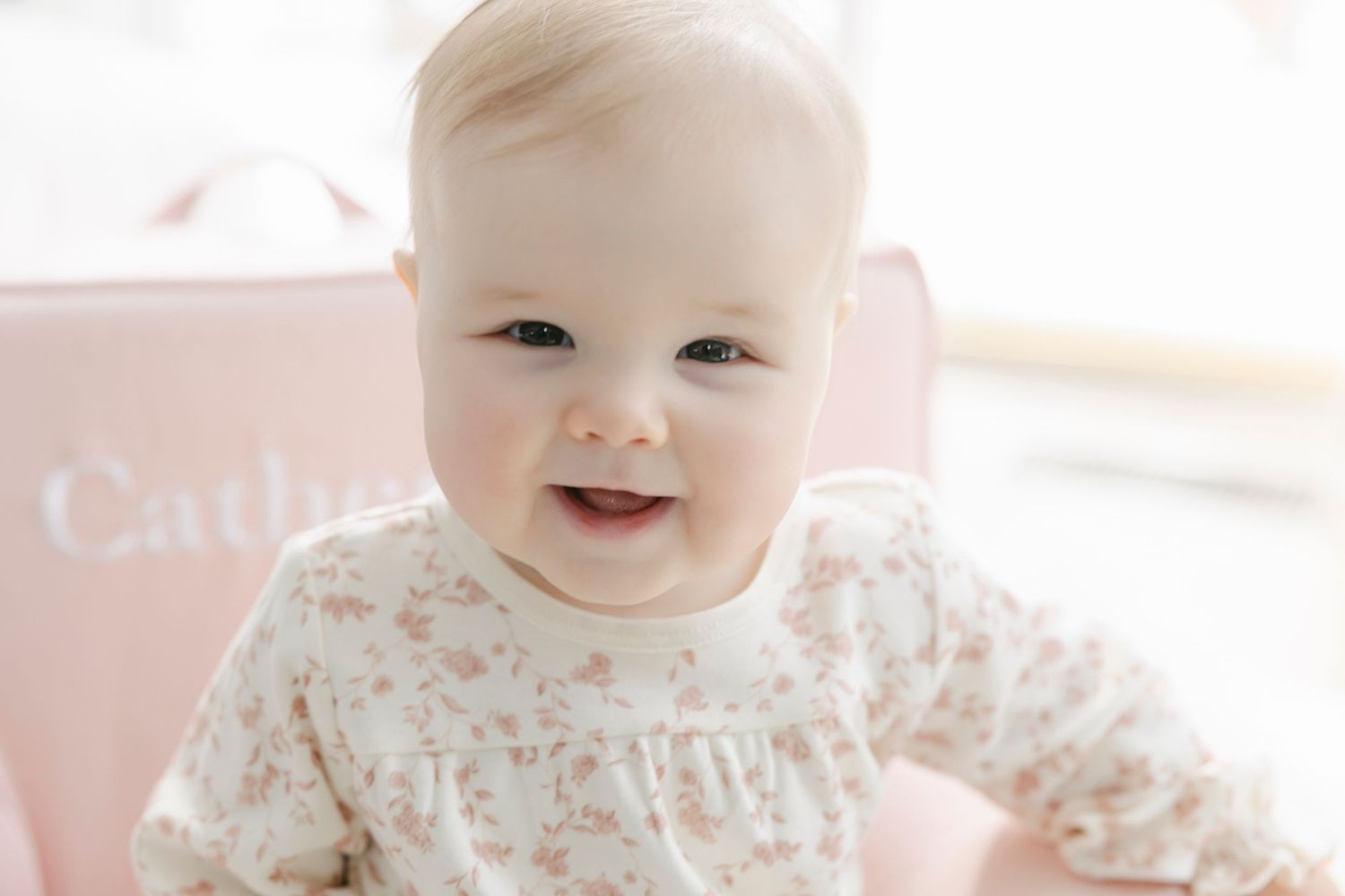 A baby wearing a floral print outfit is held up while looking at the camera in a bright indoor setting.