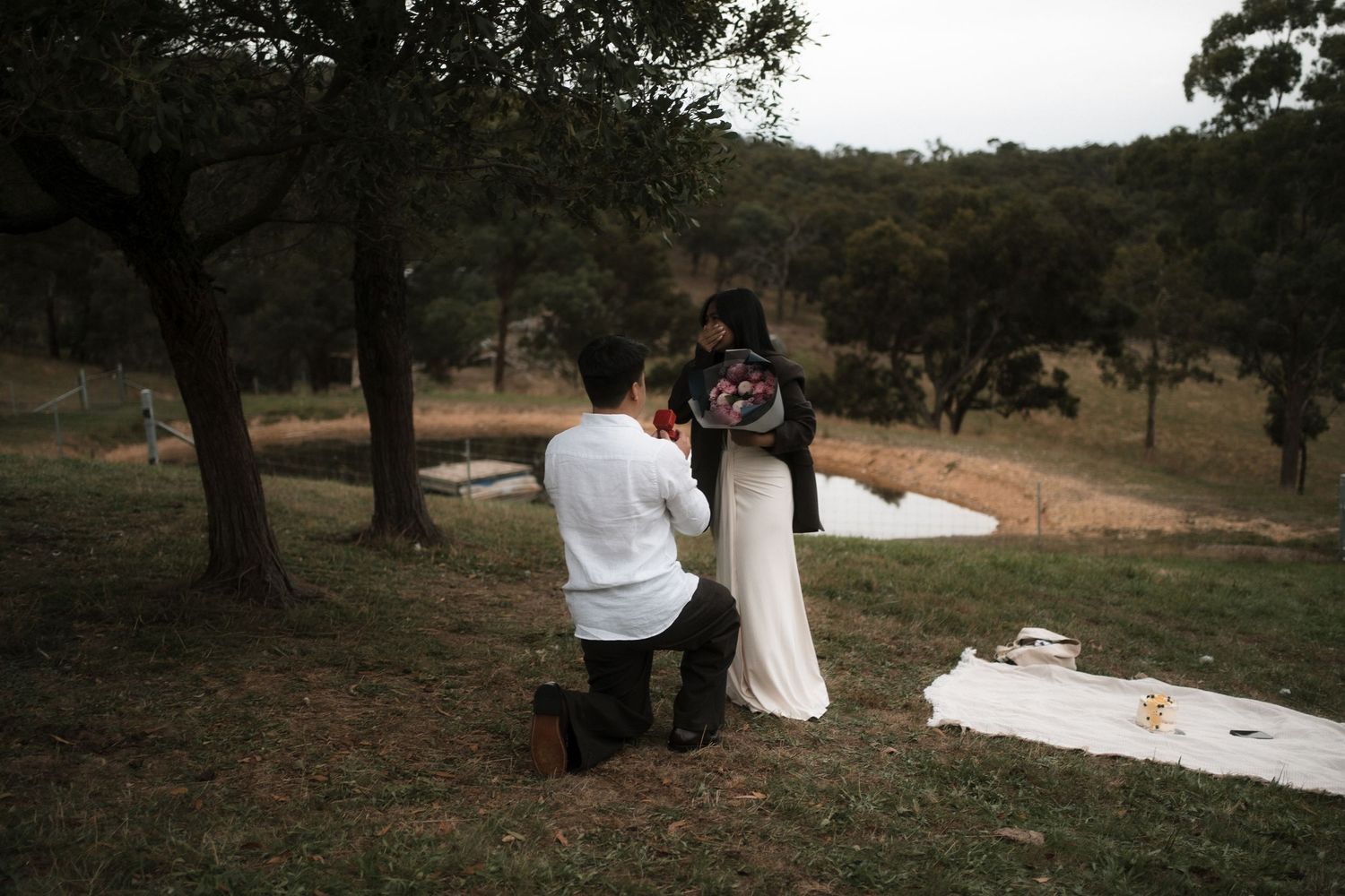 A scenic outdoor wedding ceremony takes place under tall trees with white fabric draping and wooden chairs.
