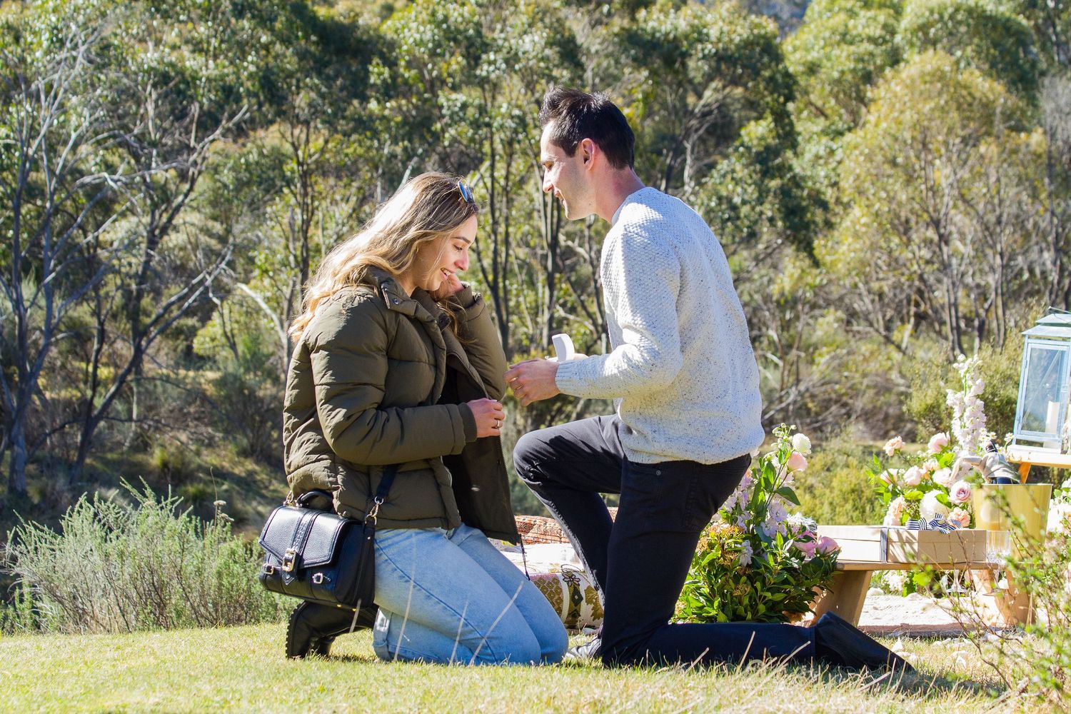 A couple shares a special moment during a proposal in a scenic outdoor garden setting with lush greenery in the background.