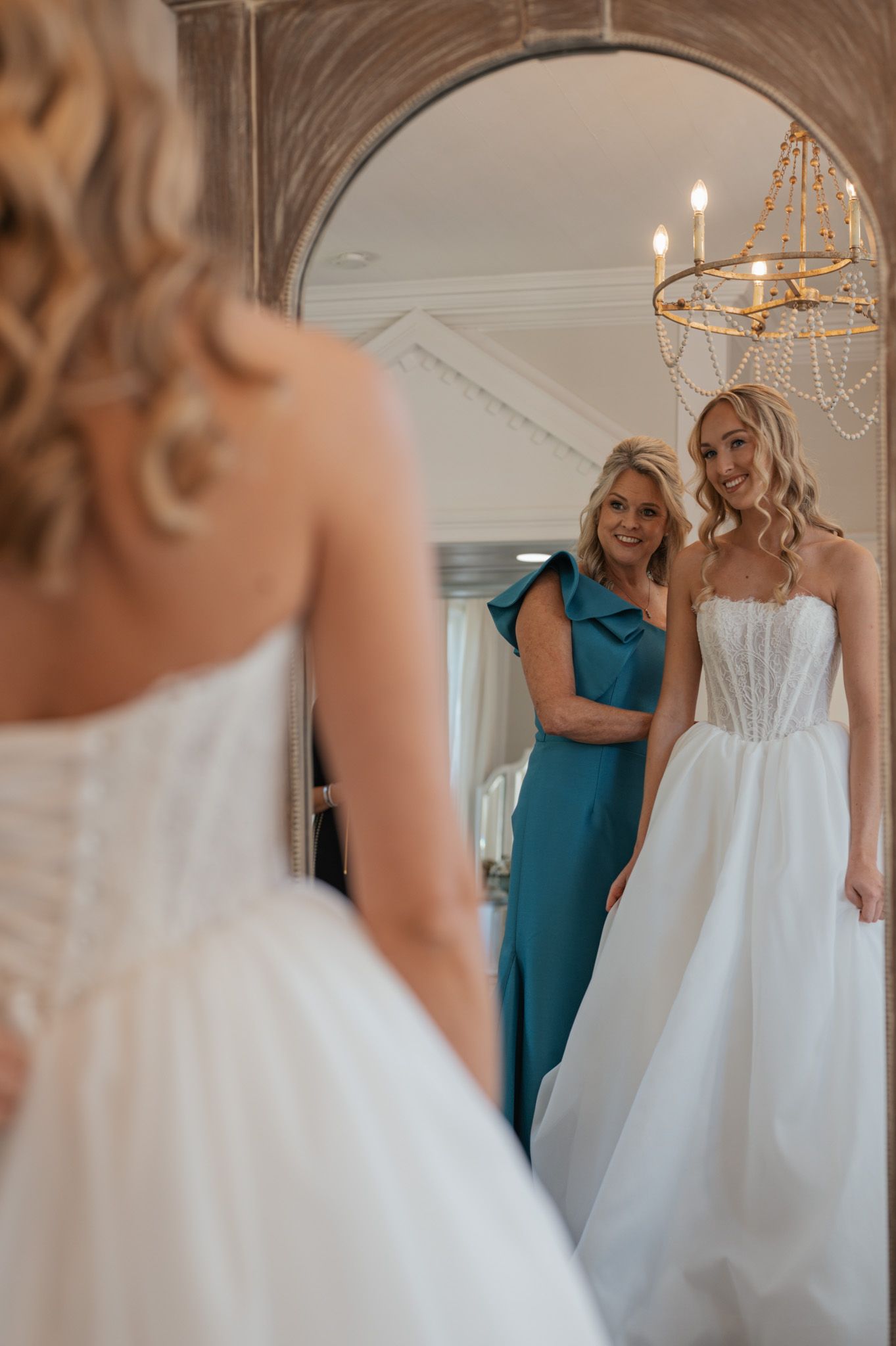 Two women share an emotional moment while getting ready in a bright room with elegant chandelier lighting.