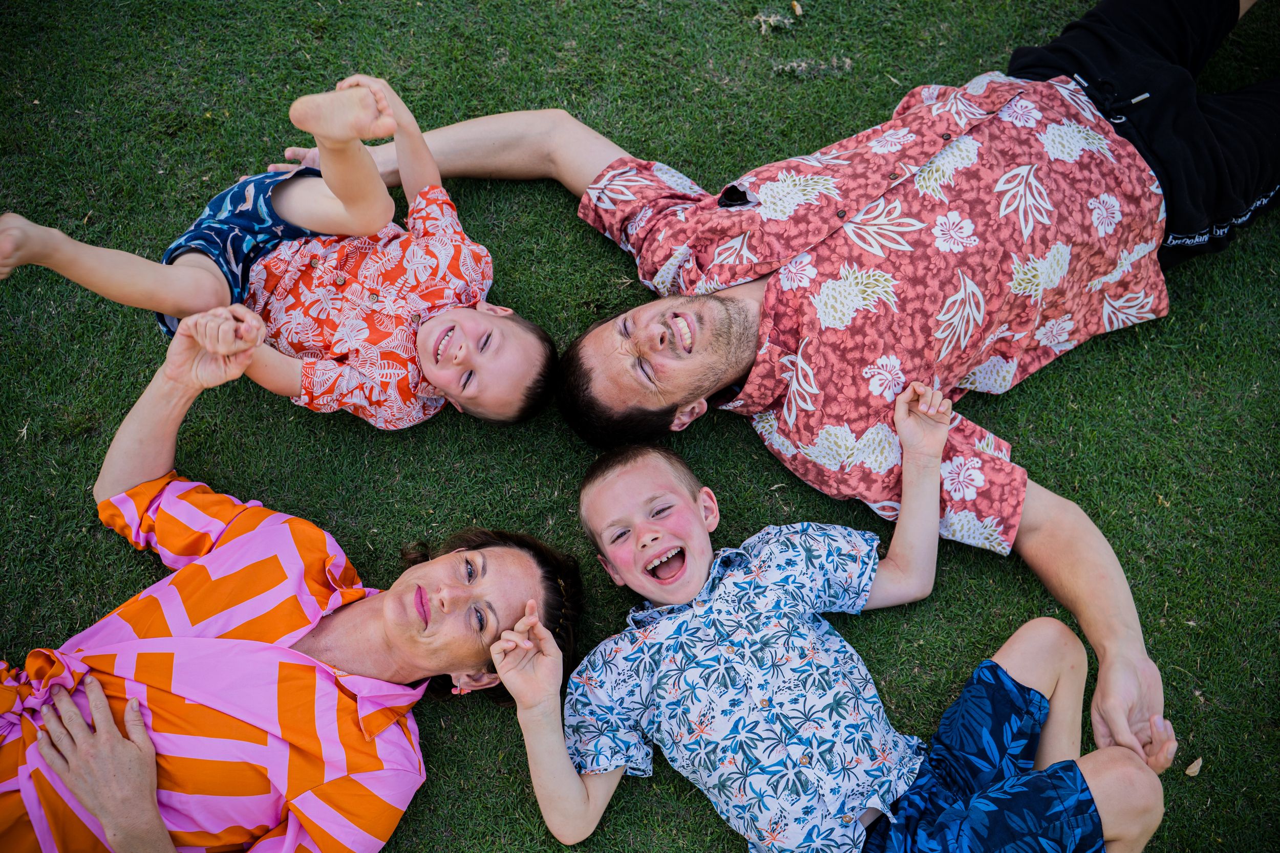 Four family members laughing and lying on green grass in colorful floral shirts, heads together in a circle.
