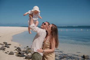 A joyful couple on a sunny beach lifting a baby in a white hat, with clear blue water in the background.