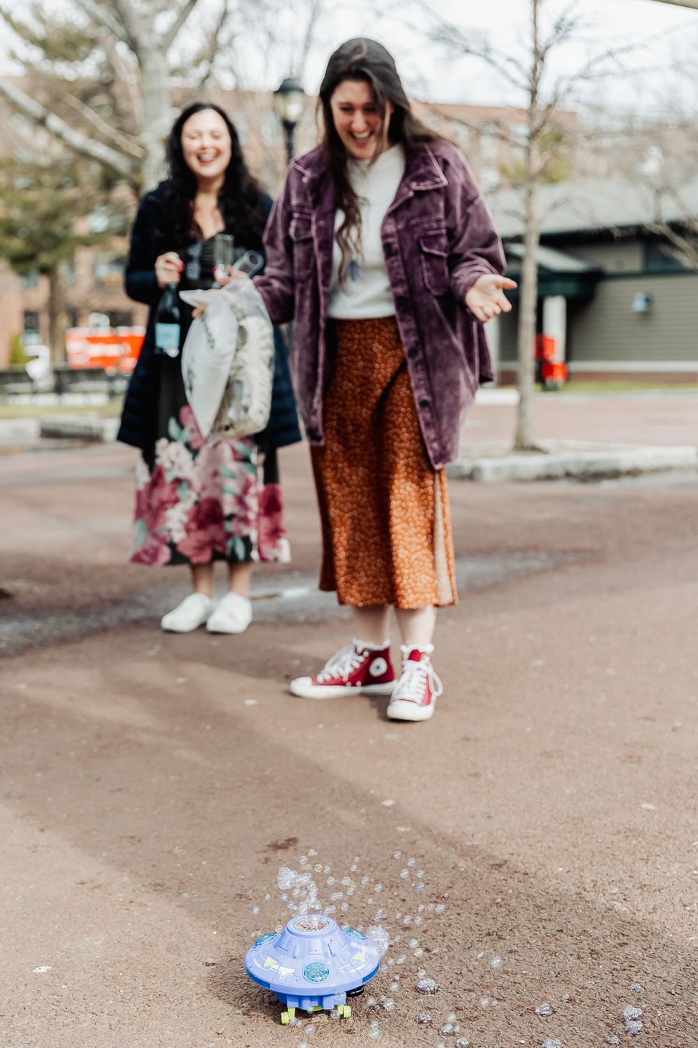 Two women laughing outdoors as a toy UFO shoots water on the pavement in a park setting.