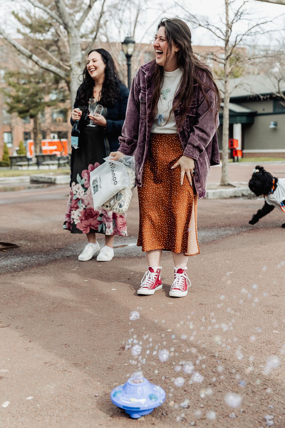 Two women laughing outdoors near a bubble machine, one in floral dress, other in orange skirt and red Converse sneakers.