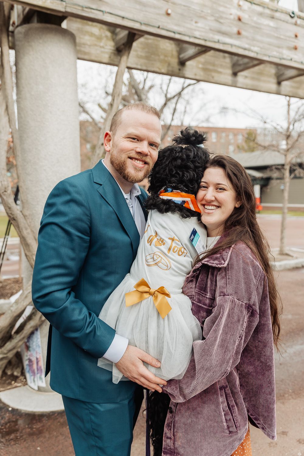 A smiling couple in a teal suit and purple jacket holds a dressed-up black poodle outdoors.