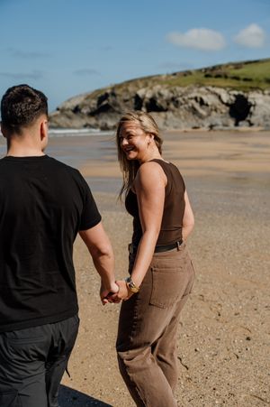 Couple holding hands on a sandy beach, woman smiling back over shoulder, coastal cliffs in background.