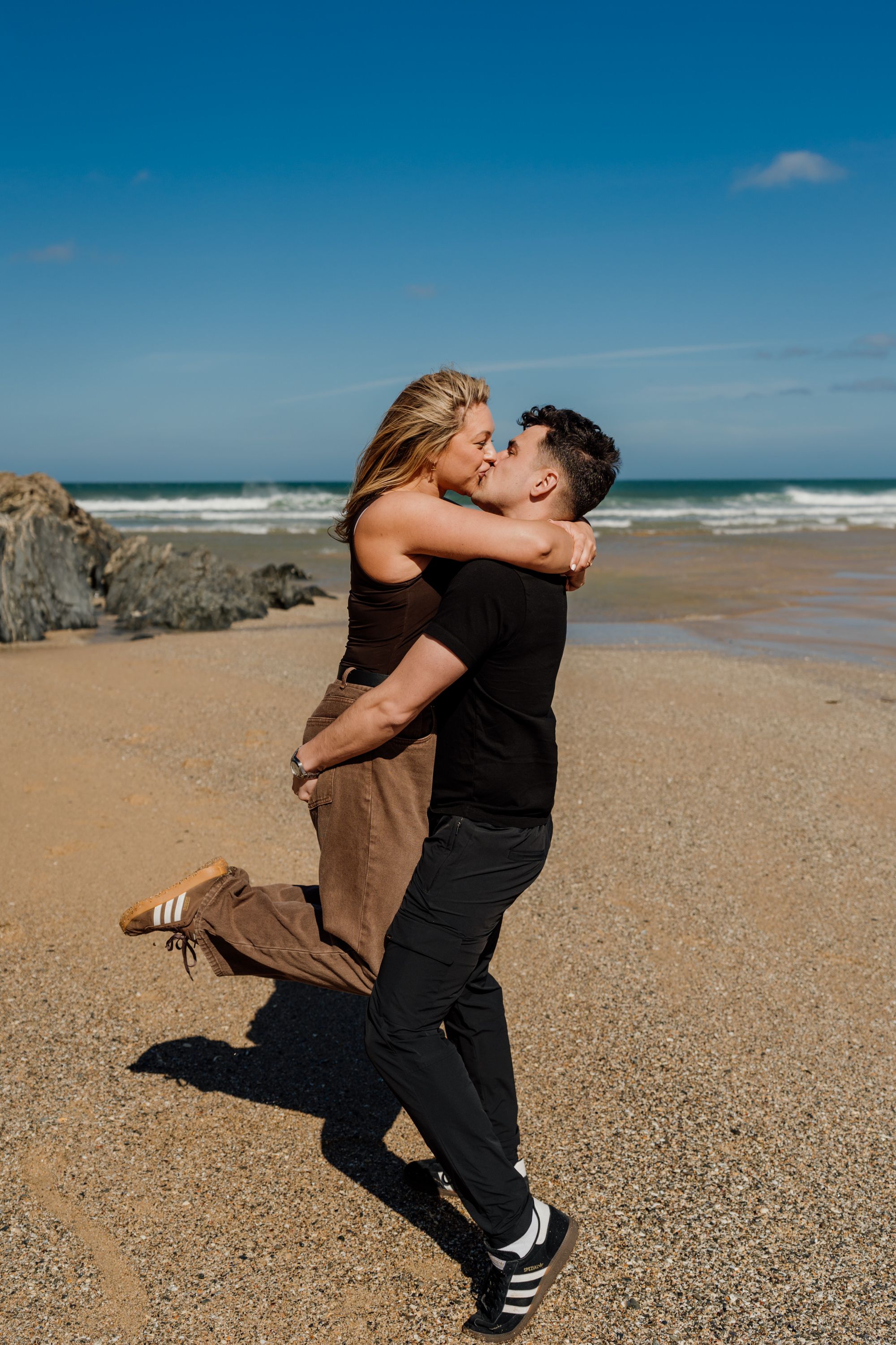Couple sharing a romantic kiss on a sunny beach, man lifting woman, ocean waves and blue sky in background.