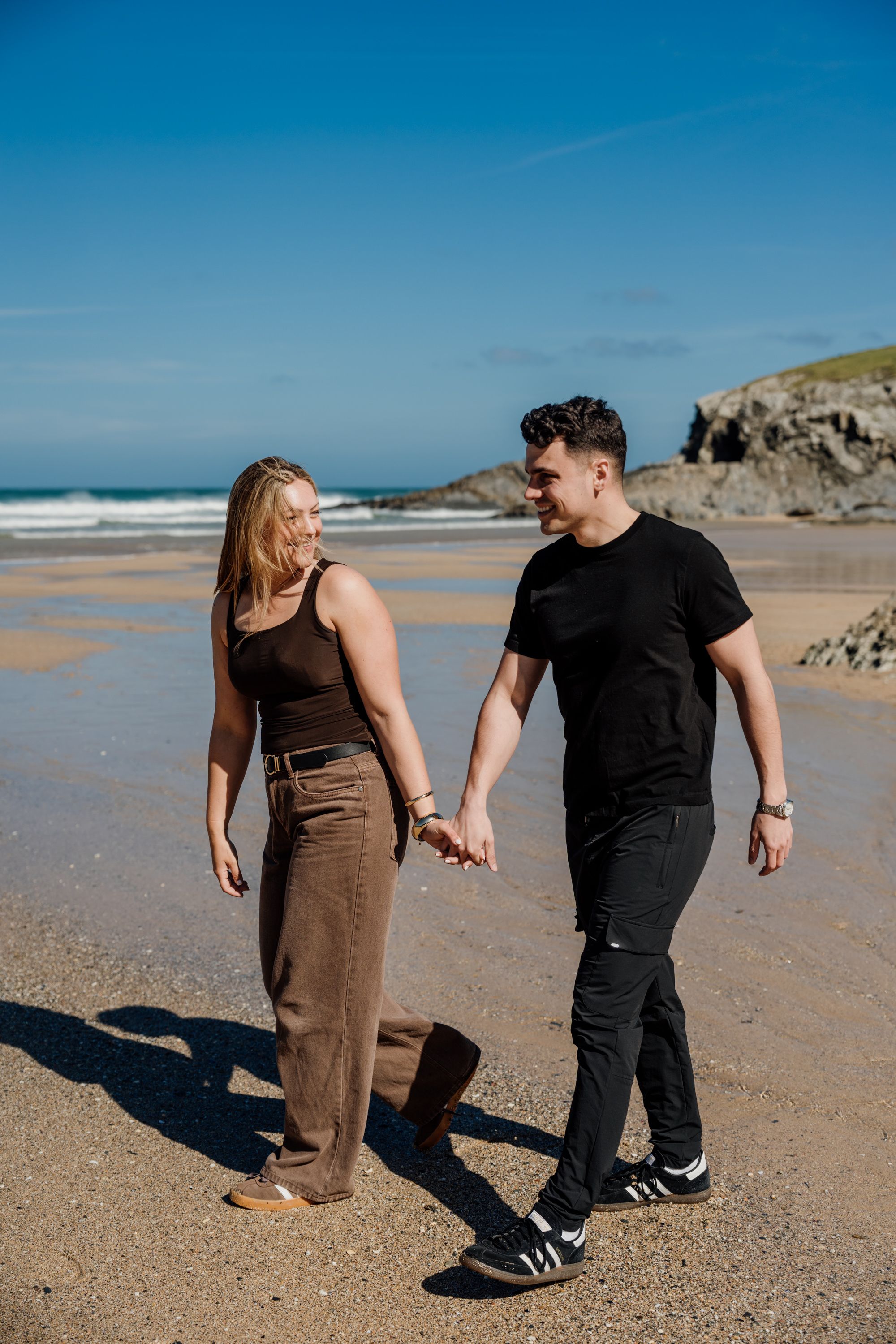 A couple holds hands and walks along a sandy beach on a sunny day with blue skies and ocean waves behind them.