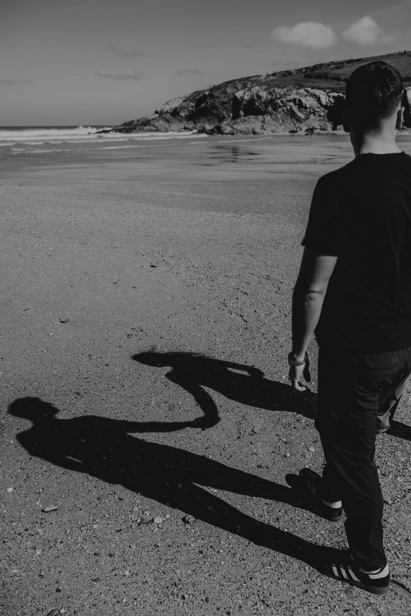 Black and white photo of a man standing on a beach casting a long shadow on the sand with rocky cliffs in background.