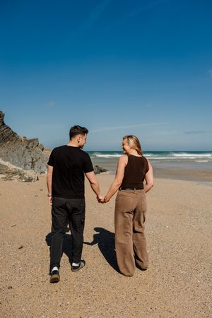 Couple holding hands walking on a sandy beach with blue sky and ocean waves in the background.