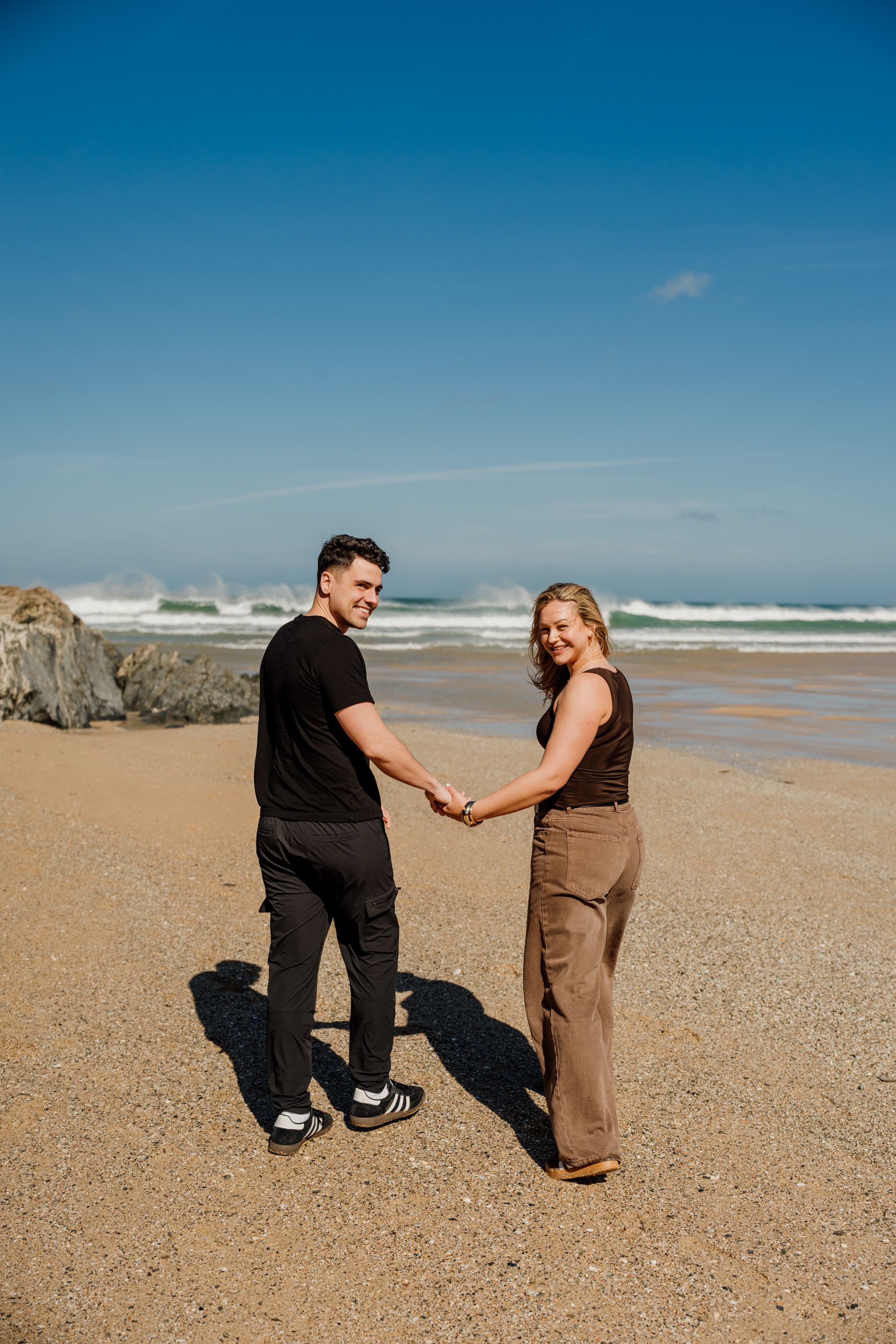 A couple holds hands and smiles on a sunny beach with ocean waves and blue sky behind them.