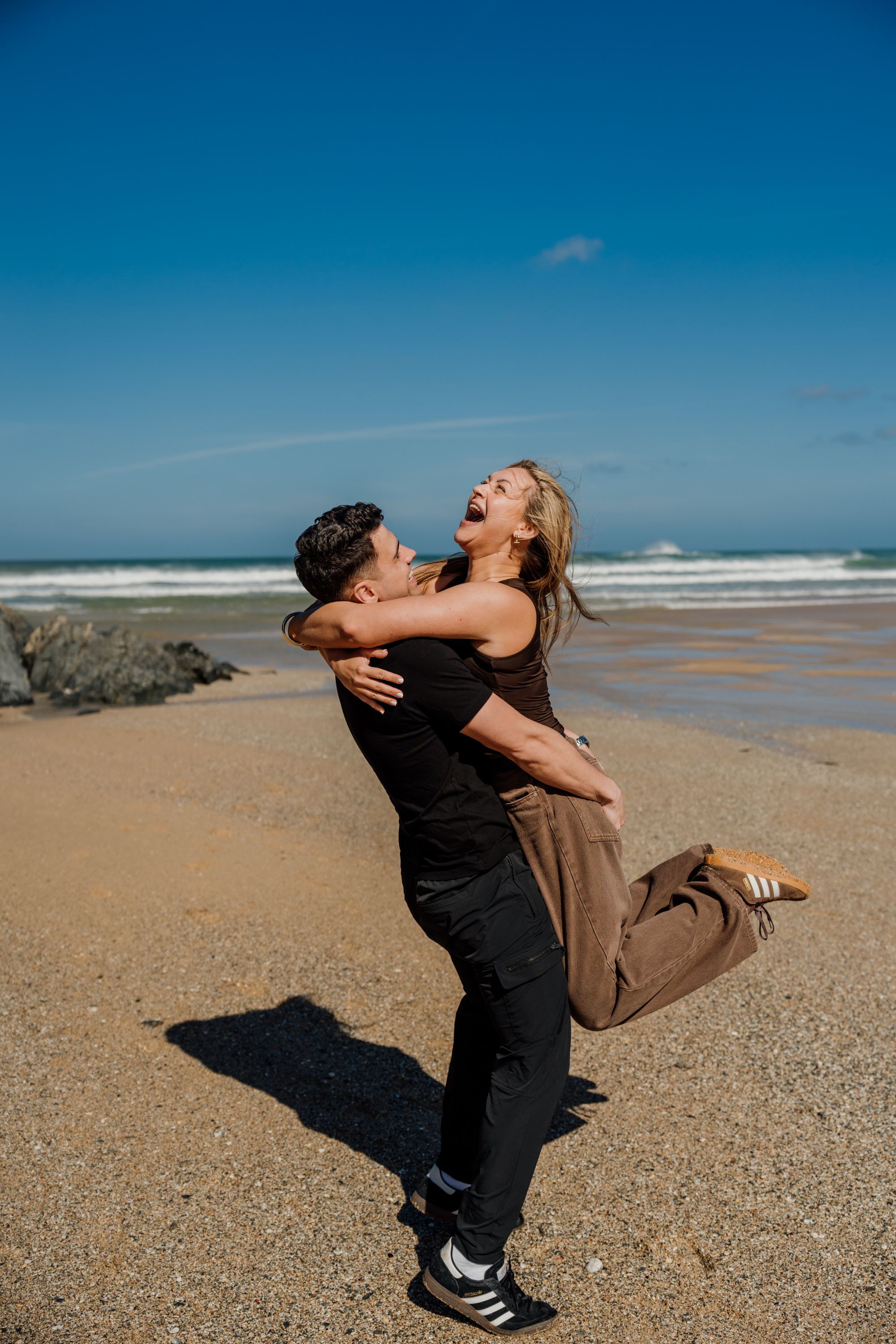 A joyful couple embracing on a sunny beach, with the man lifting the laughing woman against a blue sky backdrop.