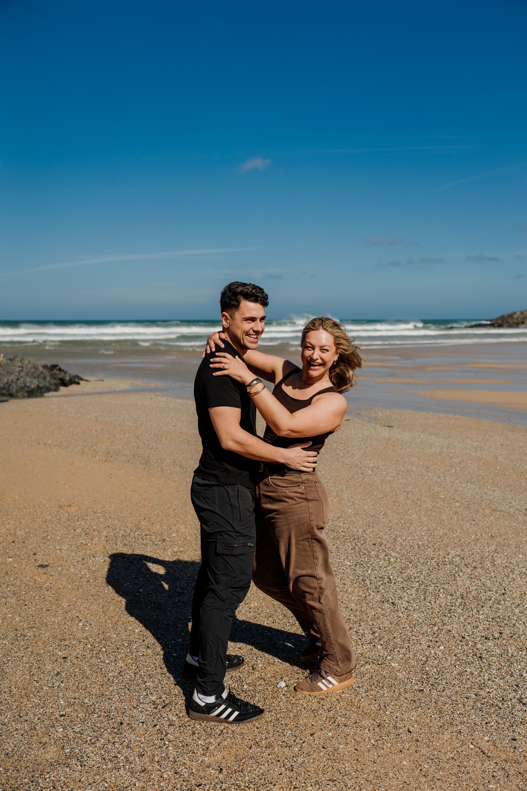 Couple embracing and laughing on a sandy beach with ocean waves and blue sky backdrop.