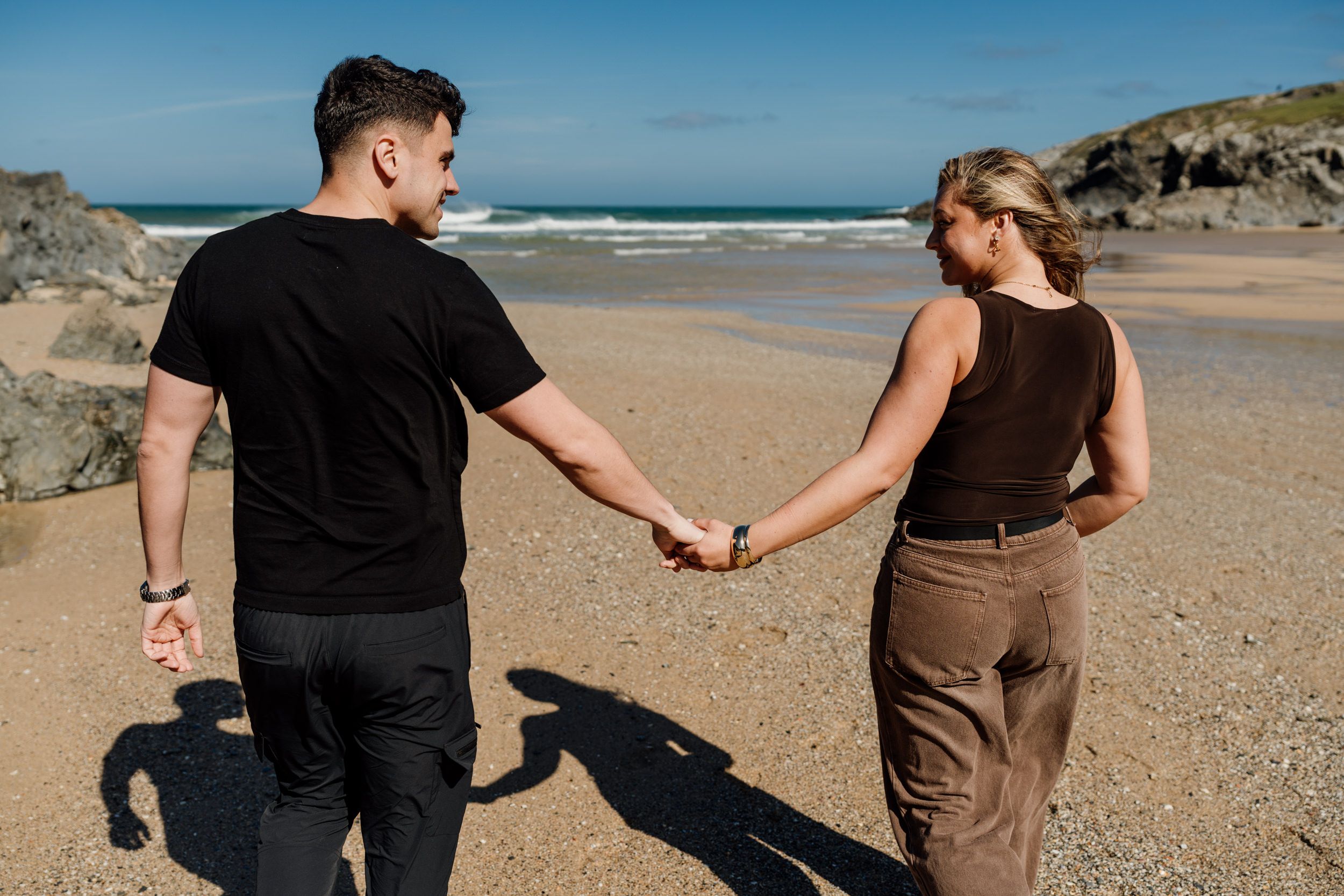 Couple holding hands walking on a sandy beach with ocean waves and rocky cliffs in the background.