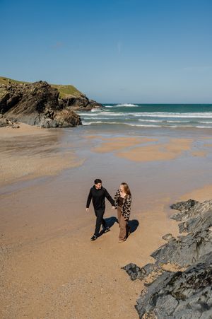 Couple holding hands walking on a sandy beach with rocky cliffs and turquoise ocean waves under a clear blue sky.