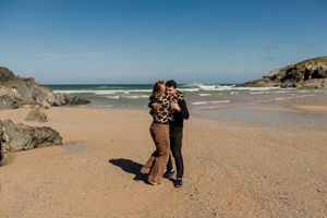 Couple embracing on a sandy beach with rocky cliffs and ocean waves under a clear blue sky.