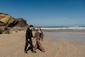 Couple holding hands walking on sandy beach with rocky cliffs and ocean waves under clear blue sky.