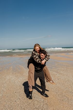 A couple laughing on a sandy beach, woman in leopard print top getting a piggyback ride from man in black.