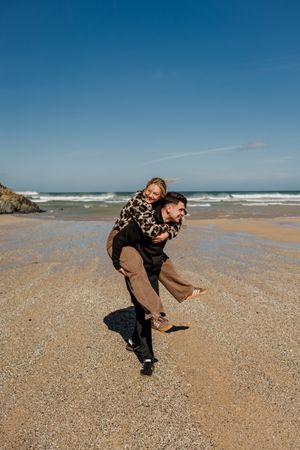 Playful couple enjoying a piggyback ride on a sunny sandy beach with ocean waves in the background.