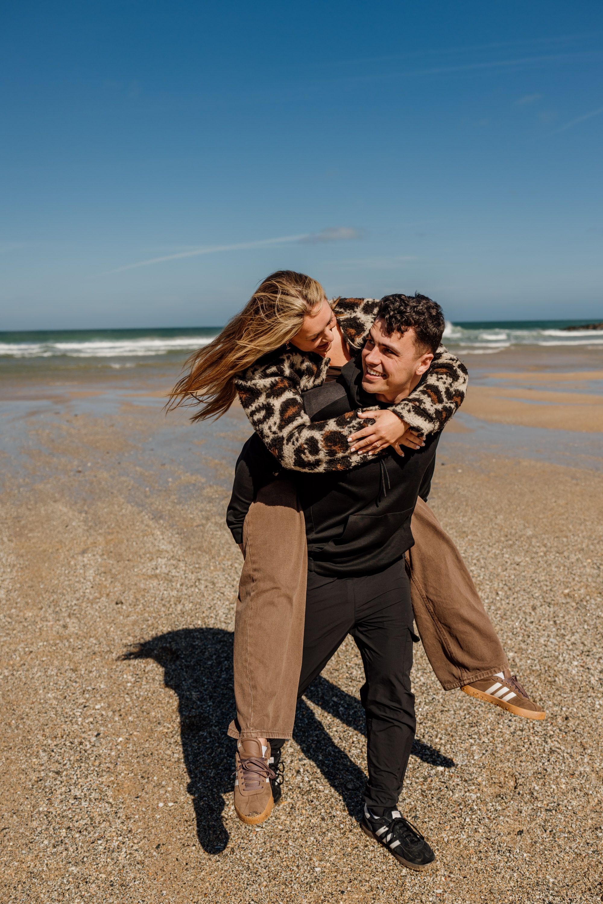 Couple laughing on a sandy beach, woman in leopard print jacket getting a piggyback ride on a sunny day.