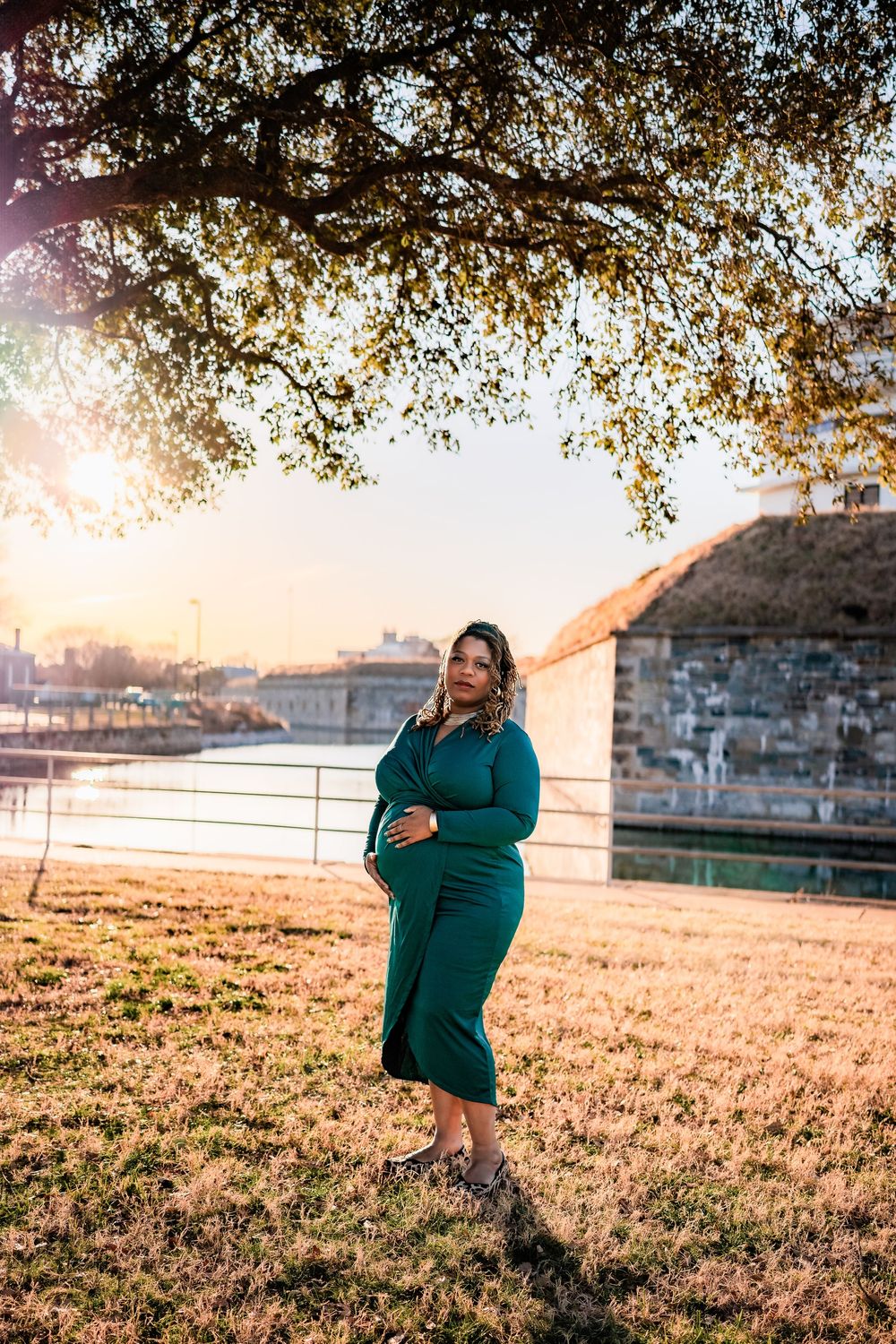 Pregnant woman in teal dress poses under tree at golden hour by riverside.