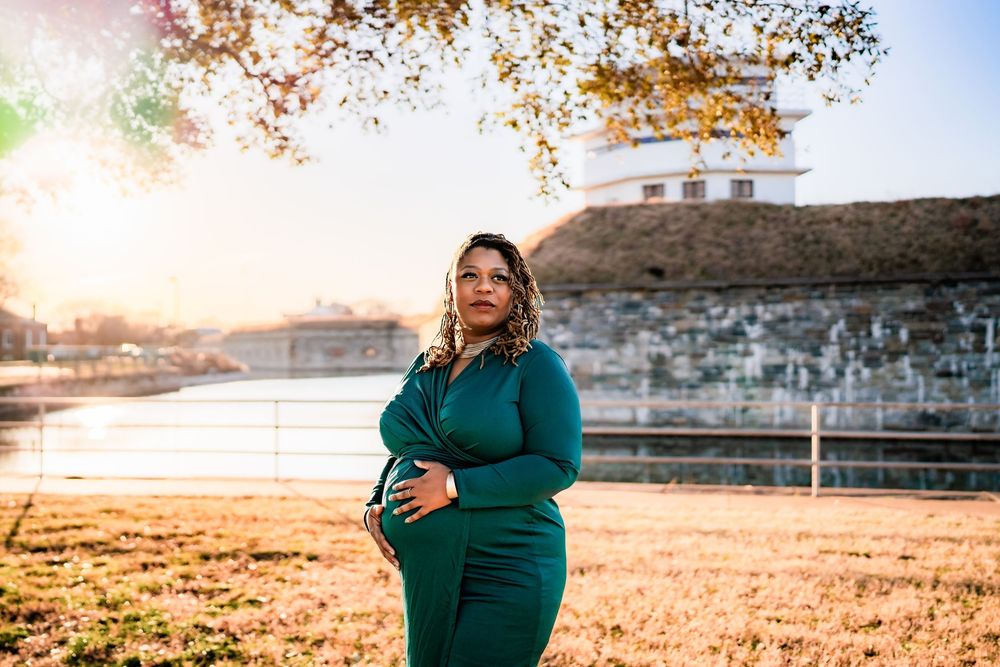 Pregnant woman in teal dress holding baby bump outdoors at golden hour near historic stone building.