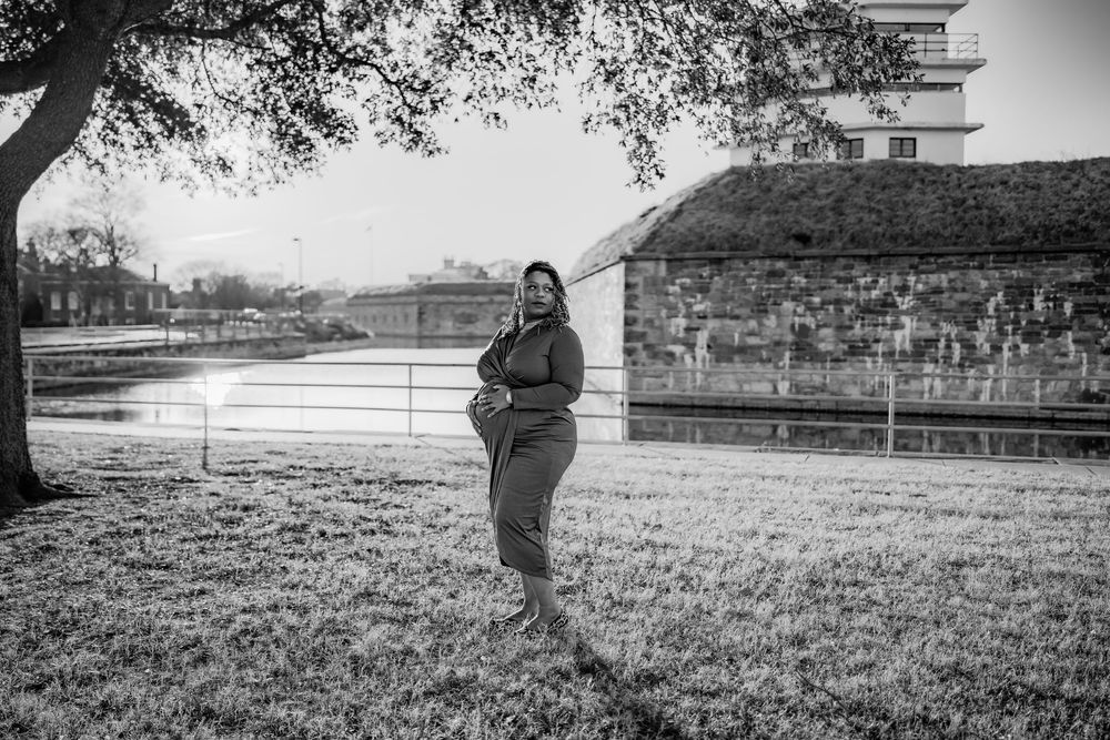 Black and white photo of a pregnant woman standing on grass near a building and pond, posing for a maternity shoot.