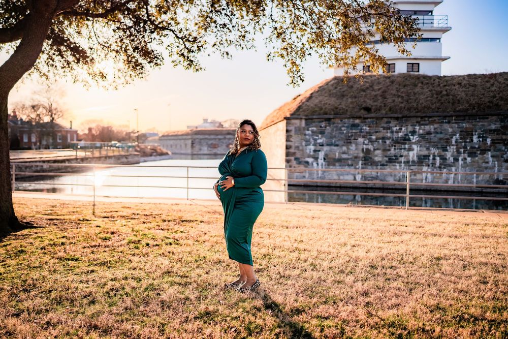 Pregnant woman in teal dress poses outdoors at golden hour near waterway and historic building.