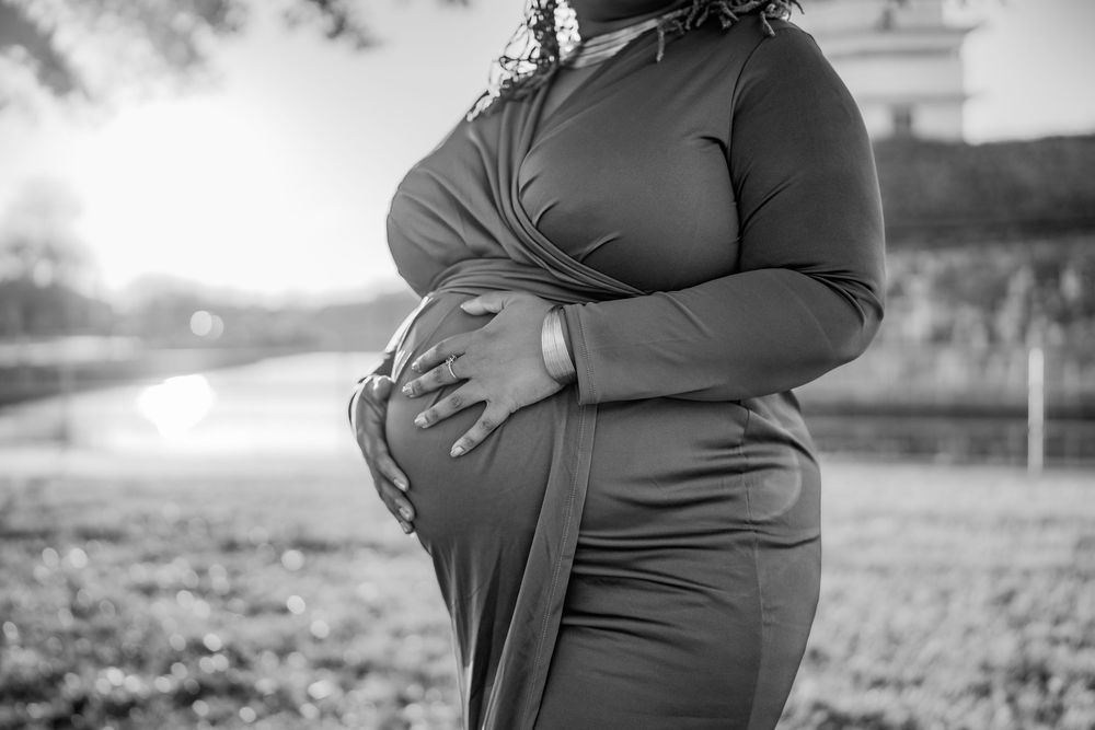 Black and white maternity photo of a pregnant woman in a fitted dress cradling her baby bump outdoors.