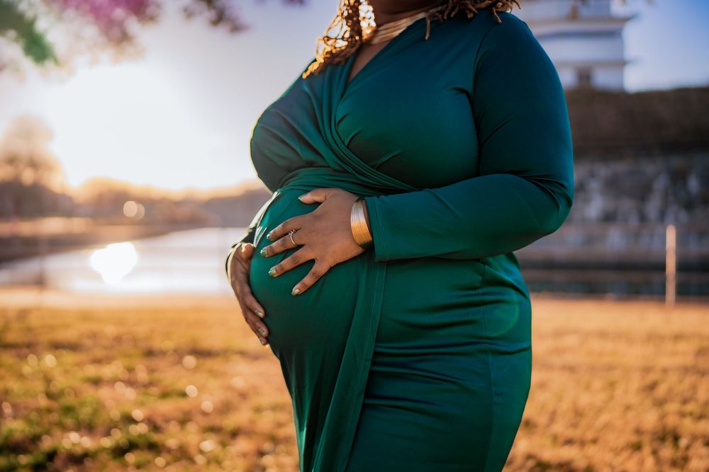 Pregnant woman in elegant green dress cradling baby bump outdoors in golden sunlight during maternity photoshoot.